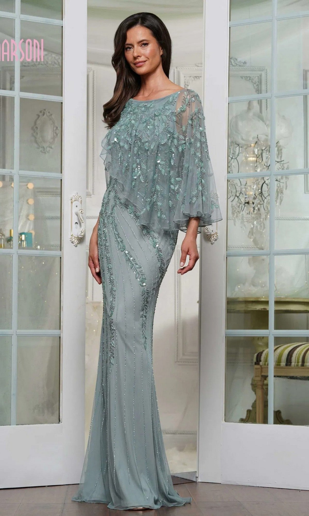 A woman in the Long Formal Dress: Marsoni MV1343, a floor-length sage gown with rhinestone embellishments, sheer beaded overlay, and floral details, stands before glass-paneled double doors in an elegant room.